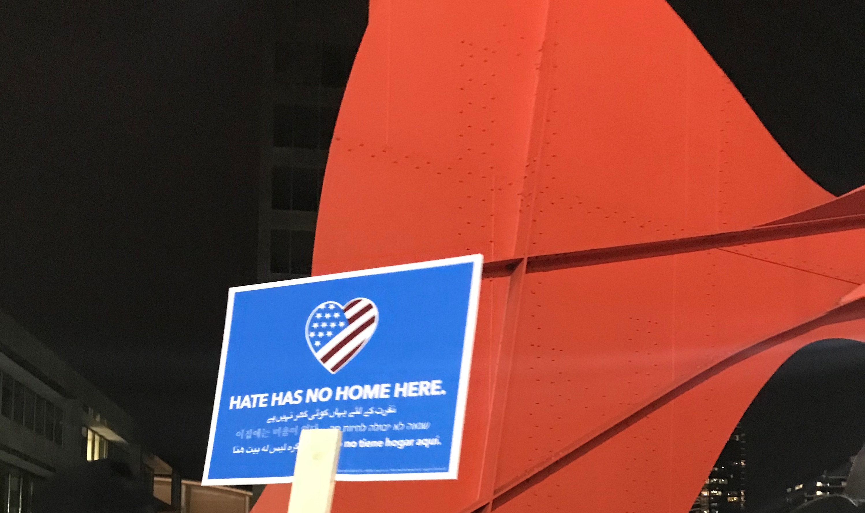 An image of the Calder sculpture in downtown Grand Rapids Michigan with a sign held up in front of it that says Hate Has No Home Here in several languages.