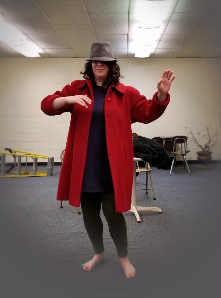 Photo of a woman with brown curly hair wearing a red coat and a fedora, dancing by herself.