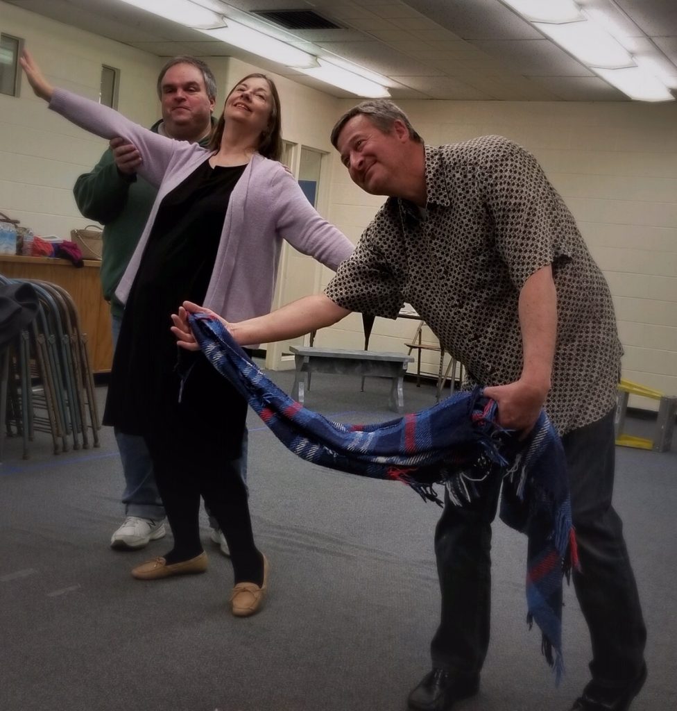 Photo of three people rehearsing a play. A man stands behind a woman, she has her arms spread wide and she is smiling. Another man is dancing with a blanket as if it is his partner.