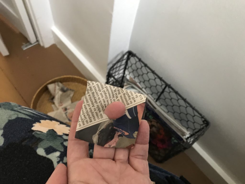 An image of a woman on the toilet with a basket of newspaper strips and a magazine caddy on the floor, her middle fingertip peeks through the middle of a folded piece of newspaper.